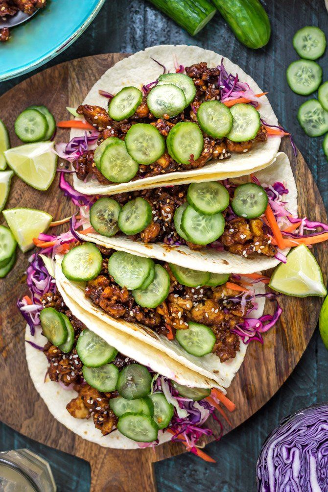 three tacos on a wooden board with cucumbers, onions, and sauce