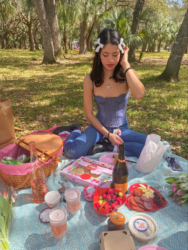 a woman sitting on the ground with food and drinks in front of her at a picnic