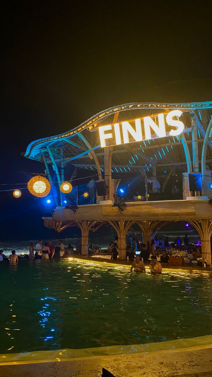 people are sitting at the edge of a swimming pool in front of an illuminated sign that reads finns