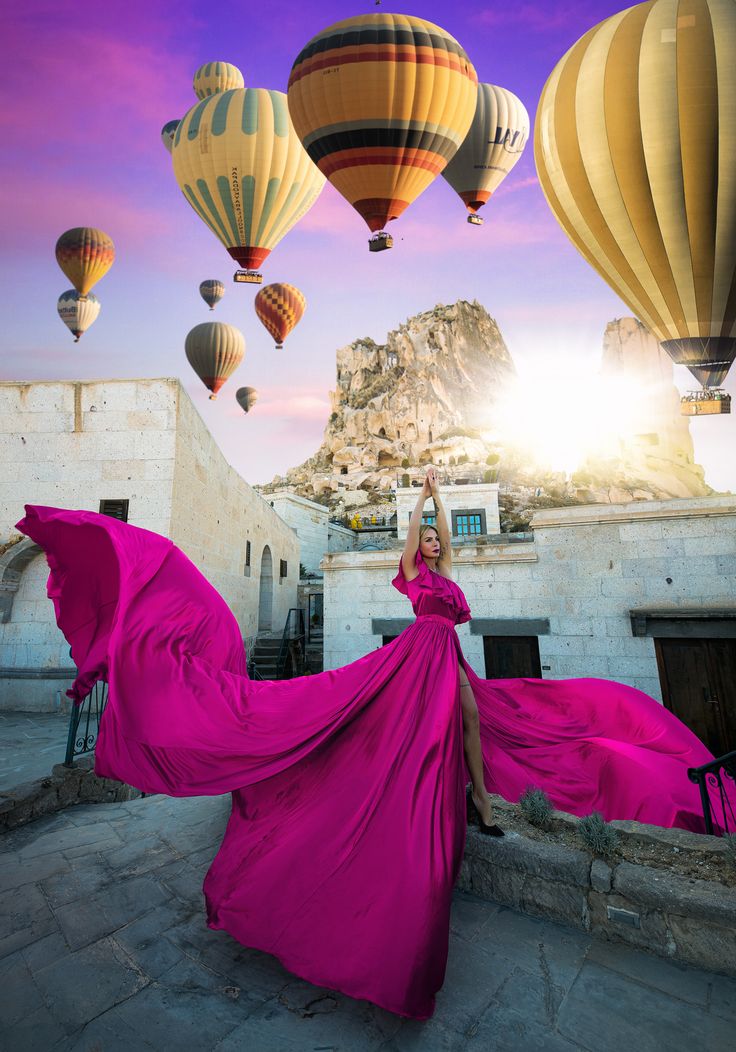 a woman in a long pink dress is posing with hot air balloons flying above her