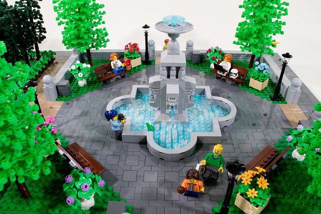 a lego model of a fountain with people sitting on benches in the center and trees around it