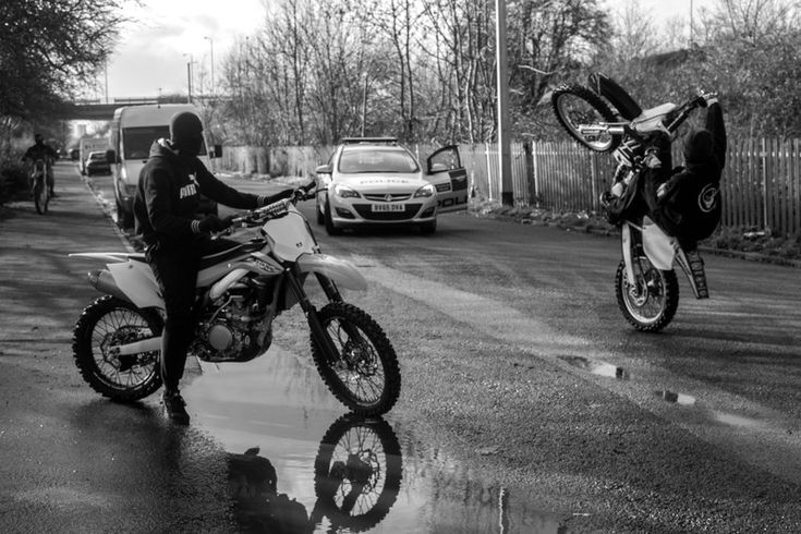 black and white photograph of two people on motorbikes in front of parked cars