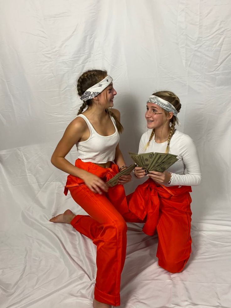 two women in red pants and white tops are sitting on a white background, one is holding money