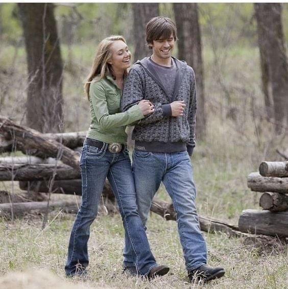 a man and woman are walking through the woods in front of some logs with trees behind them