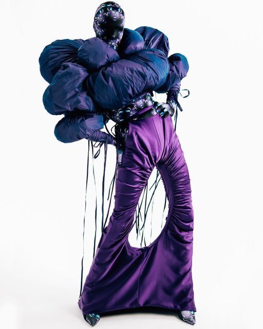 a man dressed in purple and black holding two large blue balloons on his back while standing against a white background