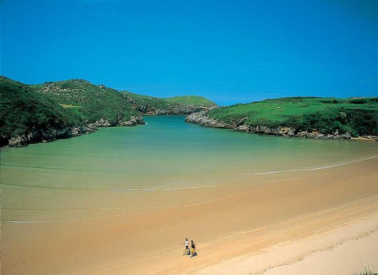 two people walking on the beach in front of some green hills and clear blue water