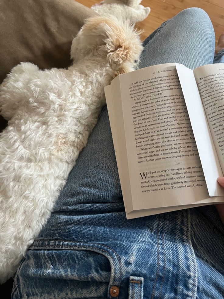 a person sitting on a couch reading a book with a dog laying next to them