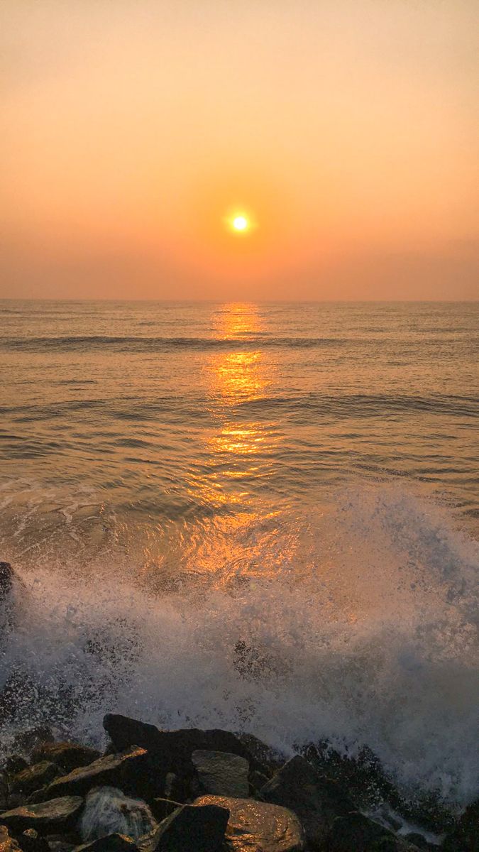 the sun is setting over the ocean as waves crash on rocks in front of it