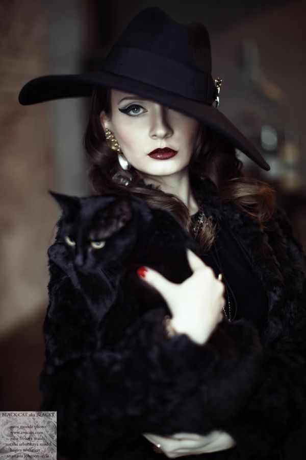 a woman in a black hat and fur coat holding a black cat with her right hand