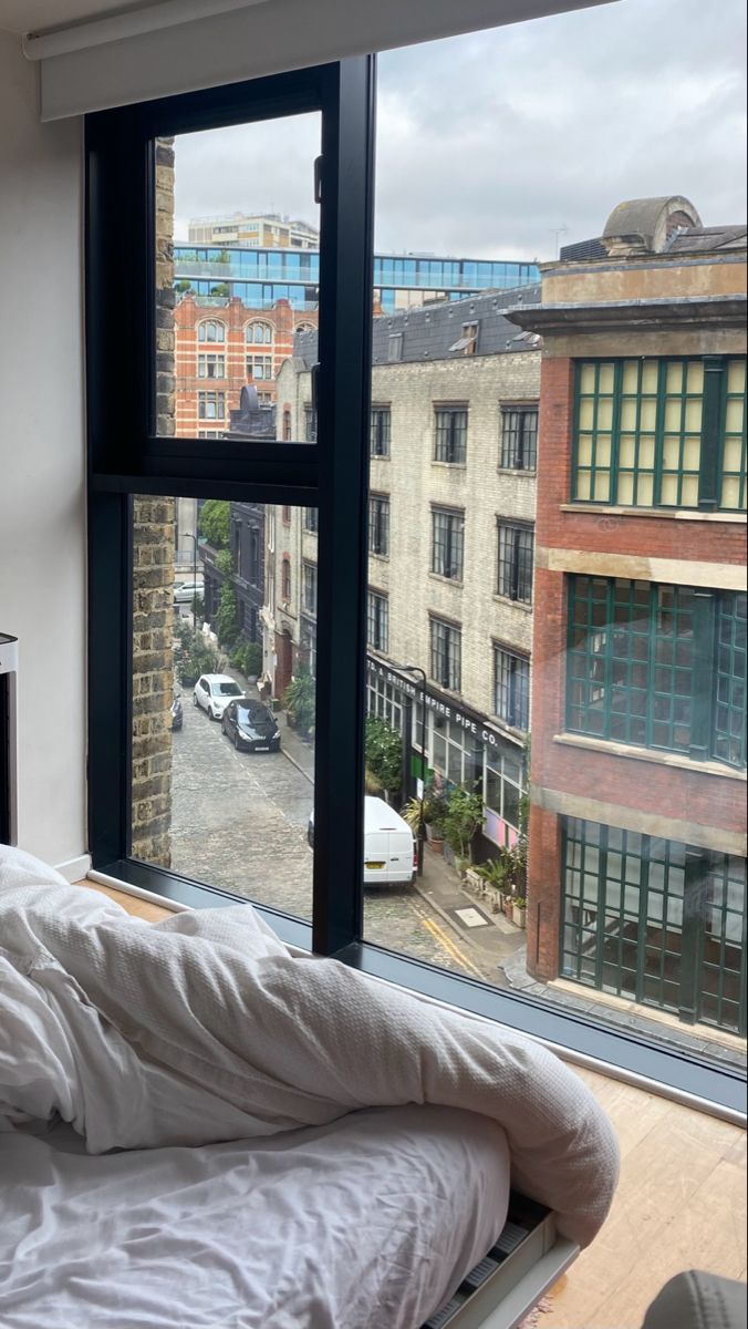 an unmade bed sitting in front of a large window with cars parked on the street outside