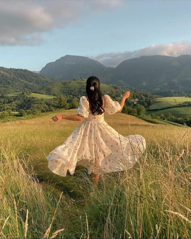 a woman in a white dress is walking through tall grass with mountains in the background