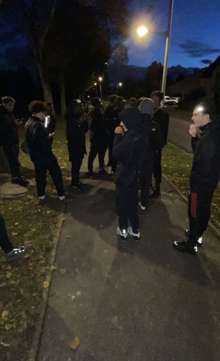 a group of people standing on the side of a road near a street light at night