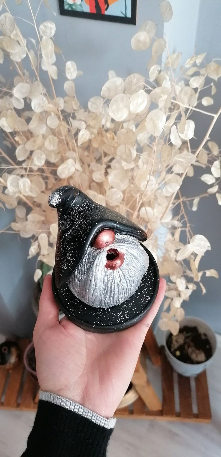 a hand holding a black vase with white flowers in it and a silver santa clause mask on top