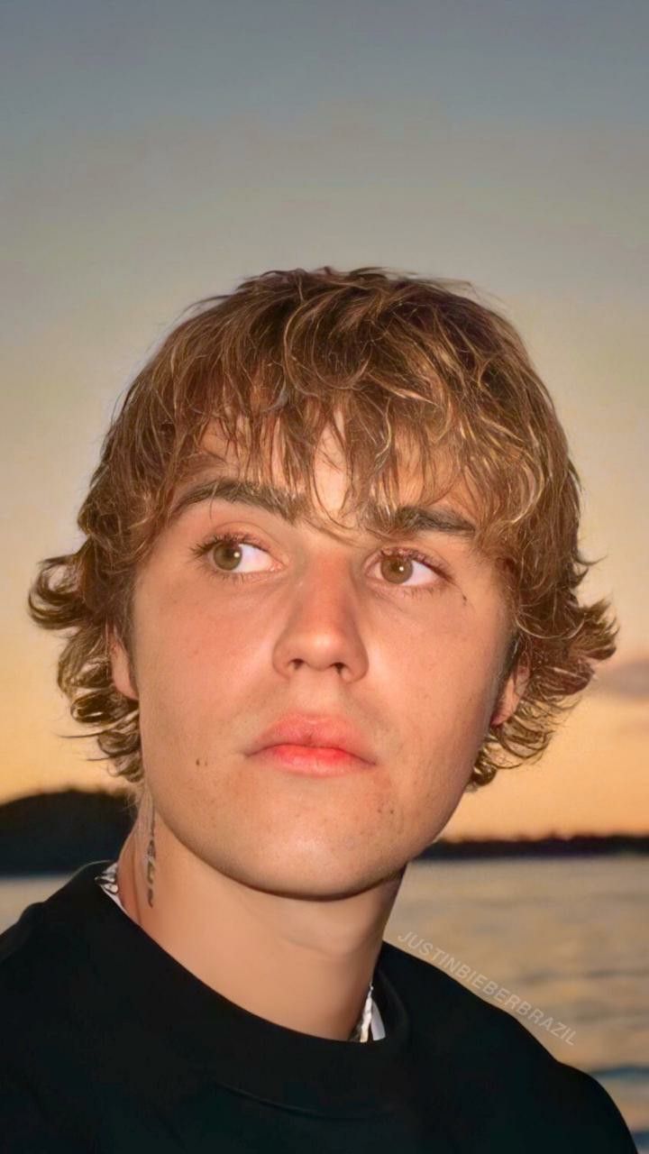 a young man standing in front of a body of water with the sun setting behind him