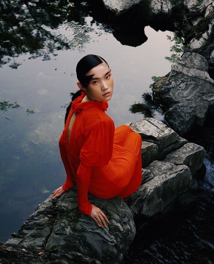 a woman in an orange dress is sitting on a rock by the water's edge