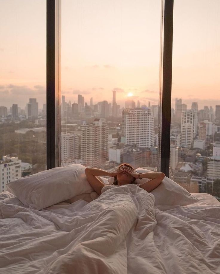 a woman laying on top of a bed in front of a window next to tall buildings