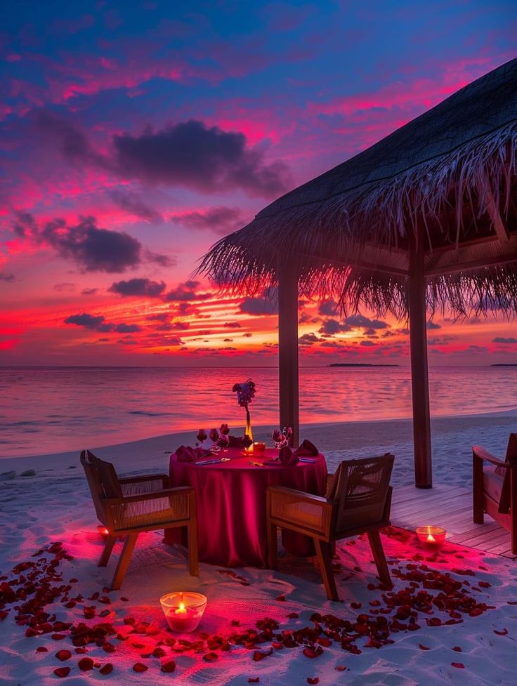 a table set up on the beach with candles and flowers in front of it at sunset