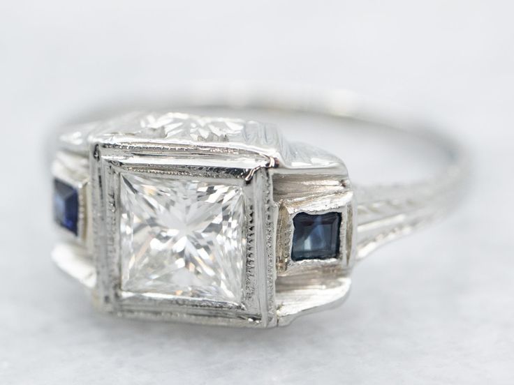 This is a lovely confection of diamond, and deep blue sapphires, set into bright crisp white 18-karat gold! This ring has all the characteristics of an Art Deco ring! The center is set with a stunning princess-cut diamond, with square-cut synthetic sapphires on either side, an iconic design of the Art Deco era!Metal: 18K White GoldGem: Diamond .47 Carats, H in Color, SI1 in Clarity Gem Measurements: 4.5 mm, Square Princess Cut Accents: 2 Synthetic Sapphires totaling .10 Carats Ring Size: 4.75Mar Princess Cut Art Deco Ring, Square Sapphire Engagement Ring, Princess Cut Diamond Ring, Diamond And Sapphire Ring, Ring Princess Cut, Deco Rings, Victorian Engagement Rings, Ring Three Stone, Diamond Ring Princess Cut