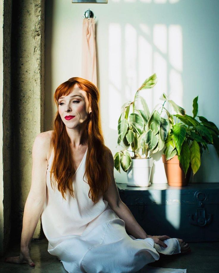 a woman with red hair sitting on the floor in front of a window and potted plant