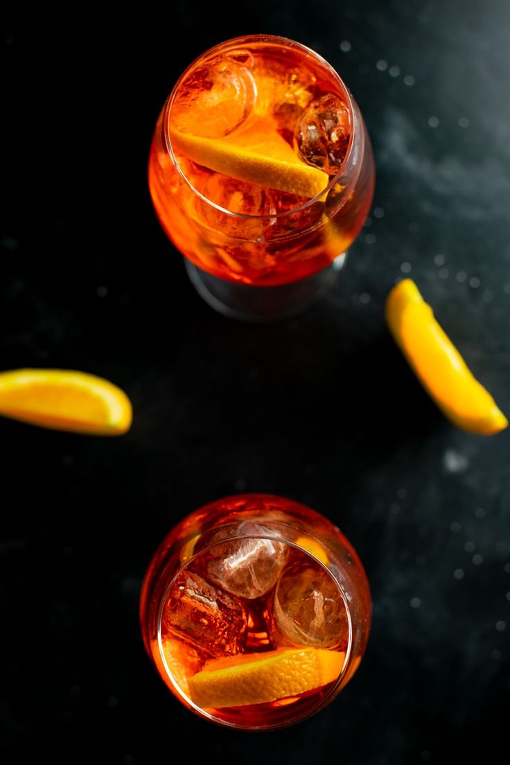 two glasses filled with ice and lemons on top of a black table next to orange slices