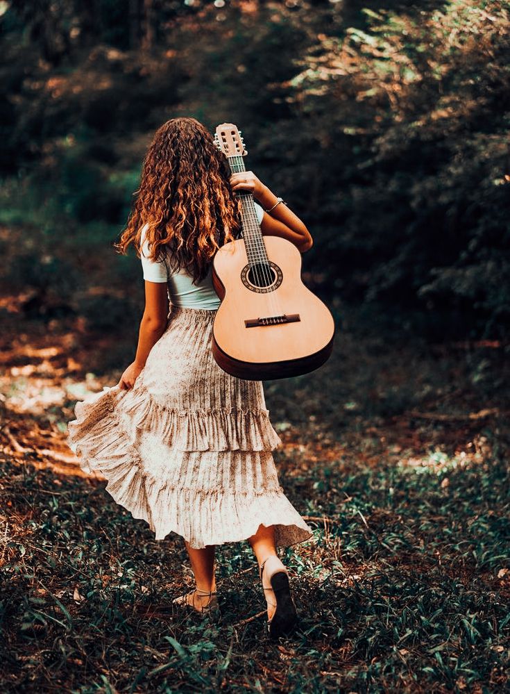 a woman in a dress is holding a guitar and walking through the woods with her back to the camera