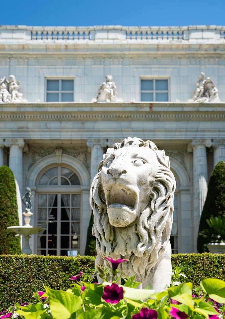 a statue of a lion in front of a building with flowers and bushes around it