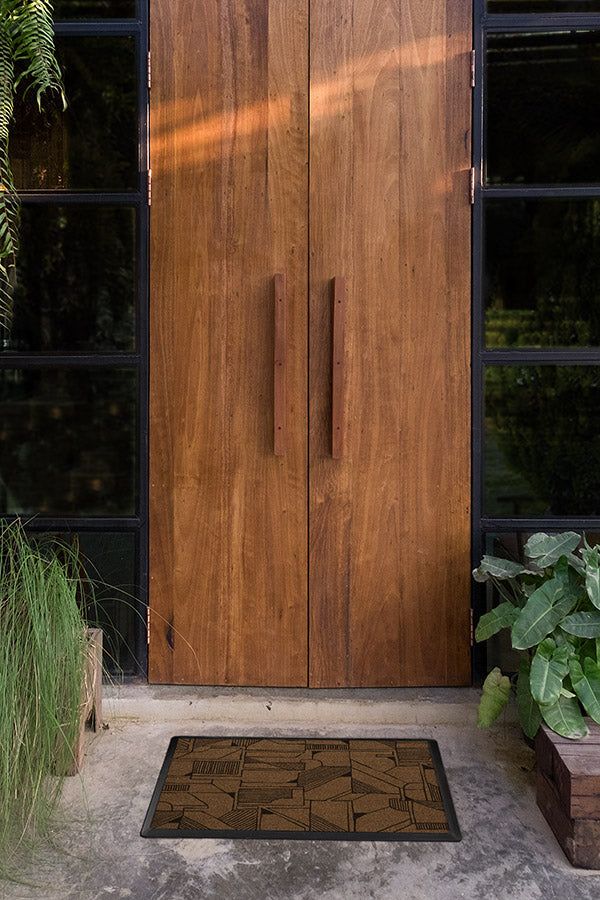 a wooden door with black windows and plants in the foreground, next to a brown mat