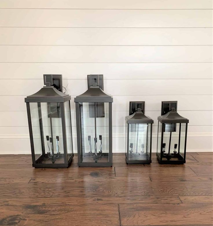 three black lanterns sitting next to each other on a wooden floor in front of a white wall