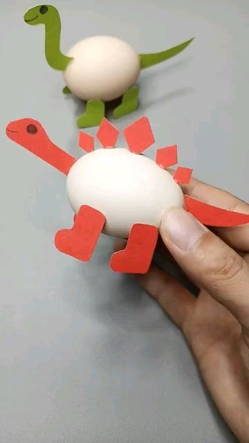 a hand is holding an egg in front of two paper dinosaurs