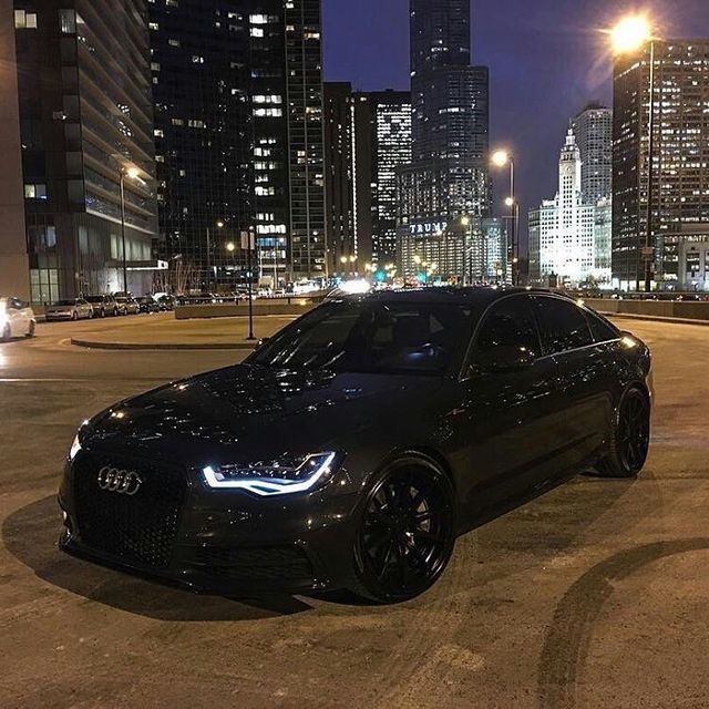 a black car parked in the middle of a parking lot with tall buildings behind it