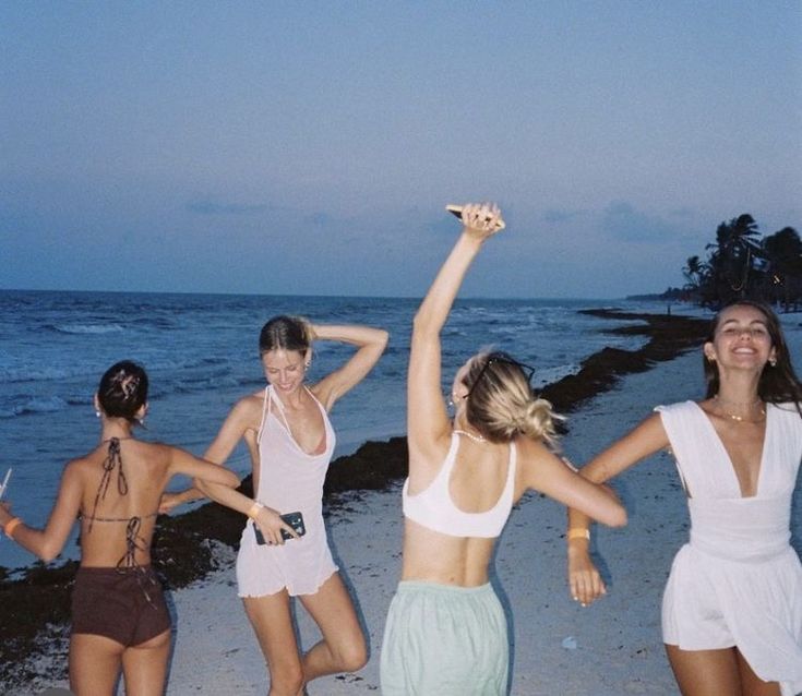 four women are dancing on the beach at night with their arms up in the air