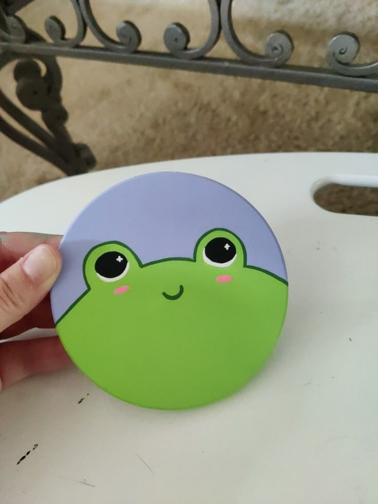 a hand holding a green frog on top of a white table next to a chair