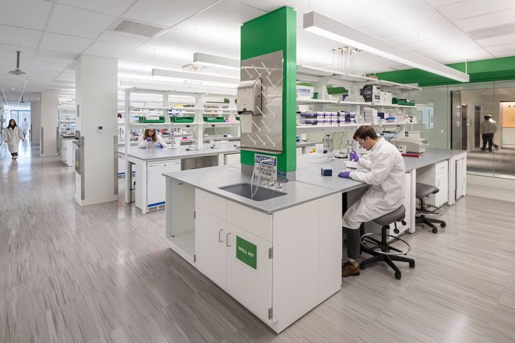 a man in white lab coat sitting at a desk with microscopes on it and people walking by