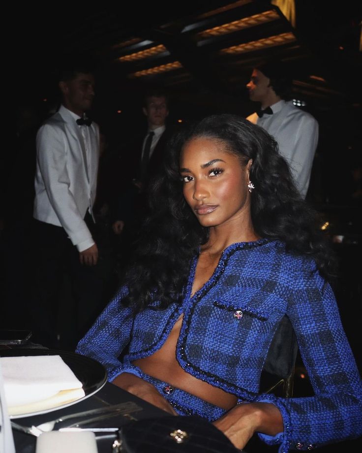 a woman sitting at a table wearing a blue top and black pants with her hands on her hips