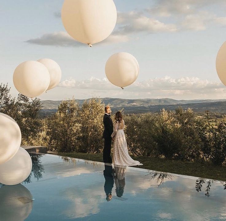 a bride and groom are standing by the pool with balloons floating in the air above them