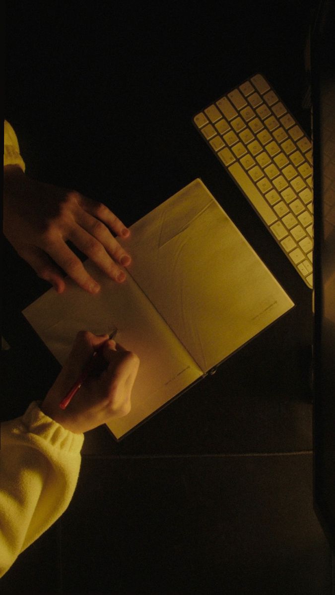 a person writing on a piece of paper next to a laptop computer and keyboard in the dark