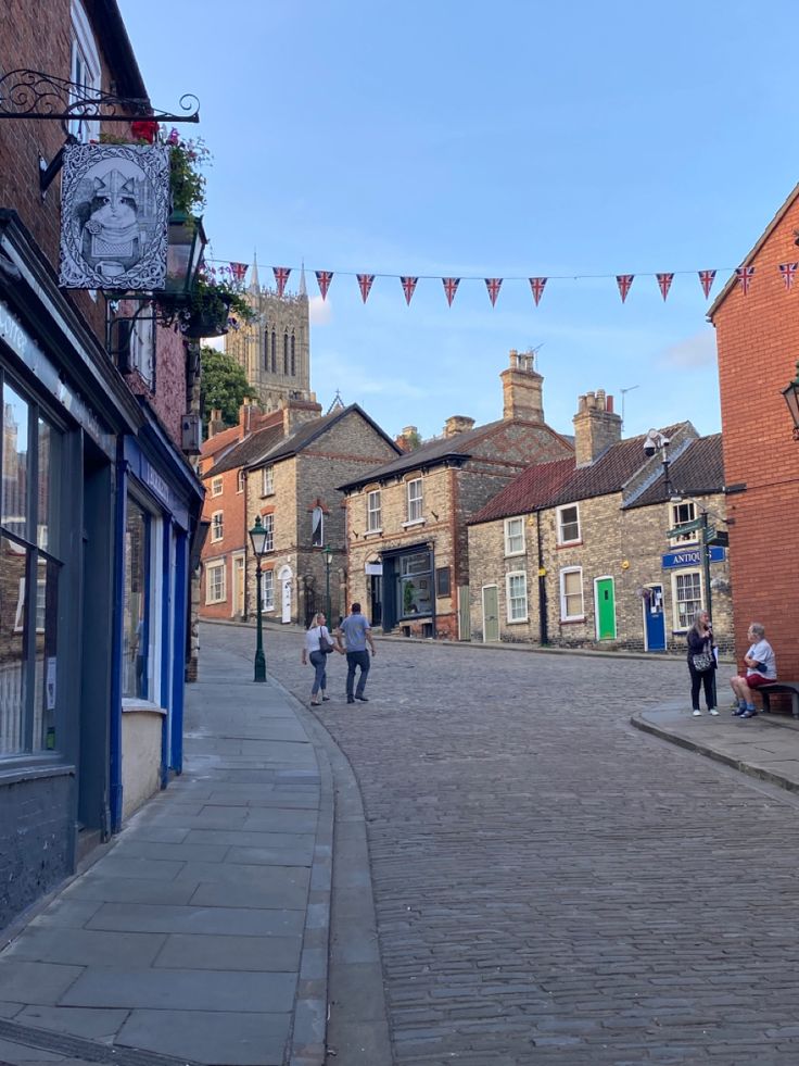 people walking down an old brick street in the middle of some small town with buildings on either side