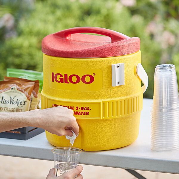 a person is pouring water into a yellow cooler on a table with other food and drinks