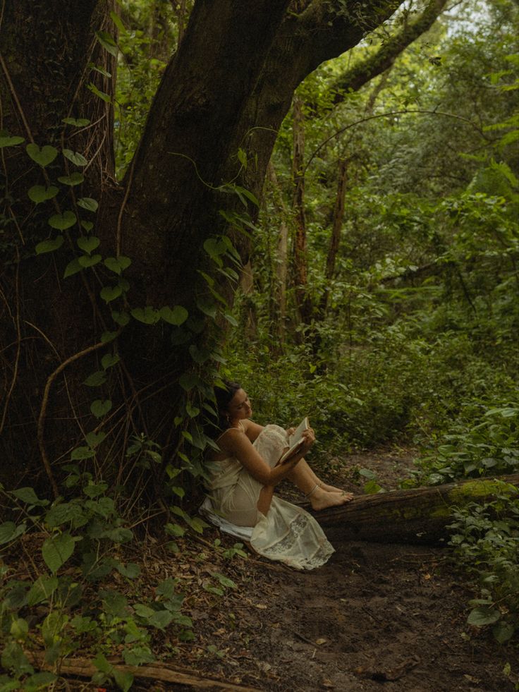 a woman sitting under a tree in the forest reading a book while wearing a white dress