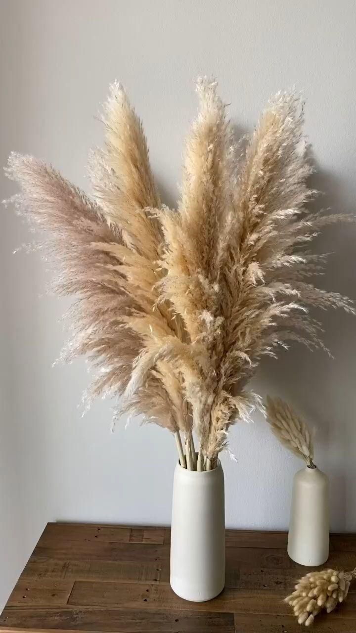 two white vases filled with dried grass on top of a wooden table