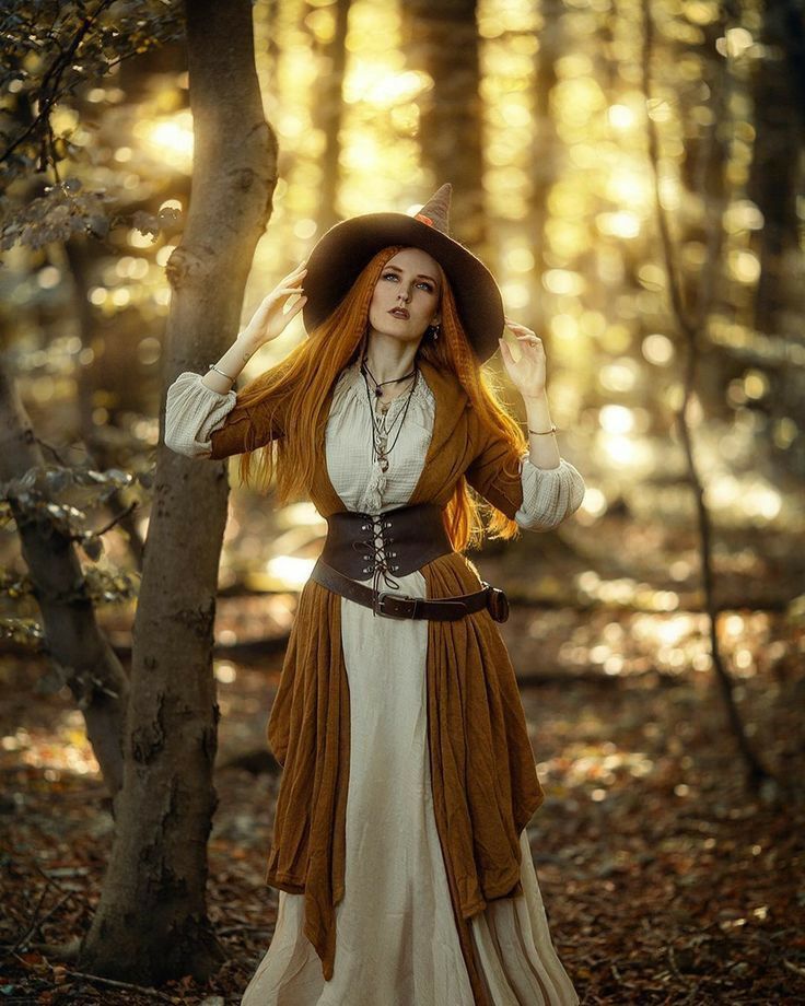 a woman with long red hair wearing a brown and white dress standing in the woods