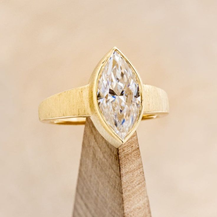 a yellow gold ring with a pear shaped diamond on the front and side, sitting on top of a wooden stand