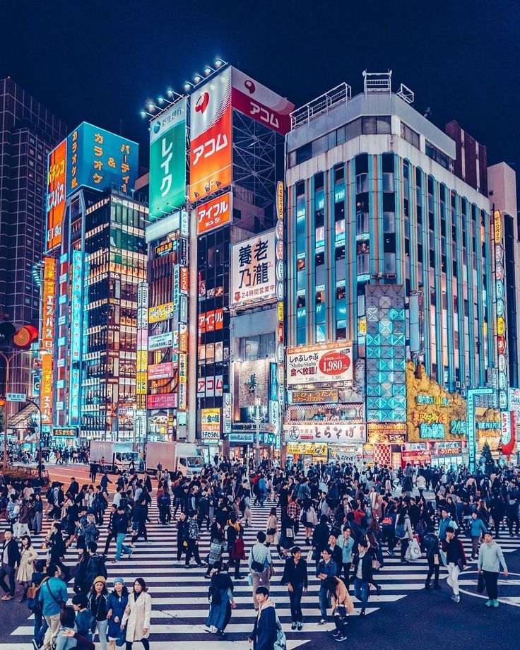 many people are crossing the street in front of some tall buildings at night with bright lights