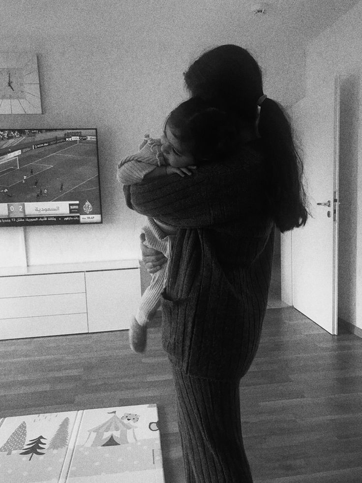 a woman holding a child in her arms while standing on the floor with a flat screen tv behind her