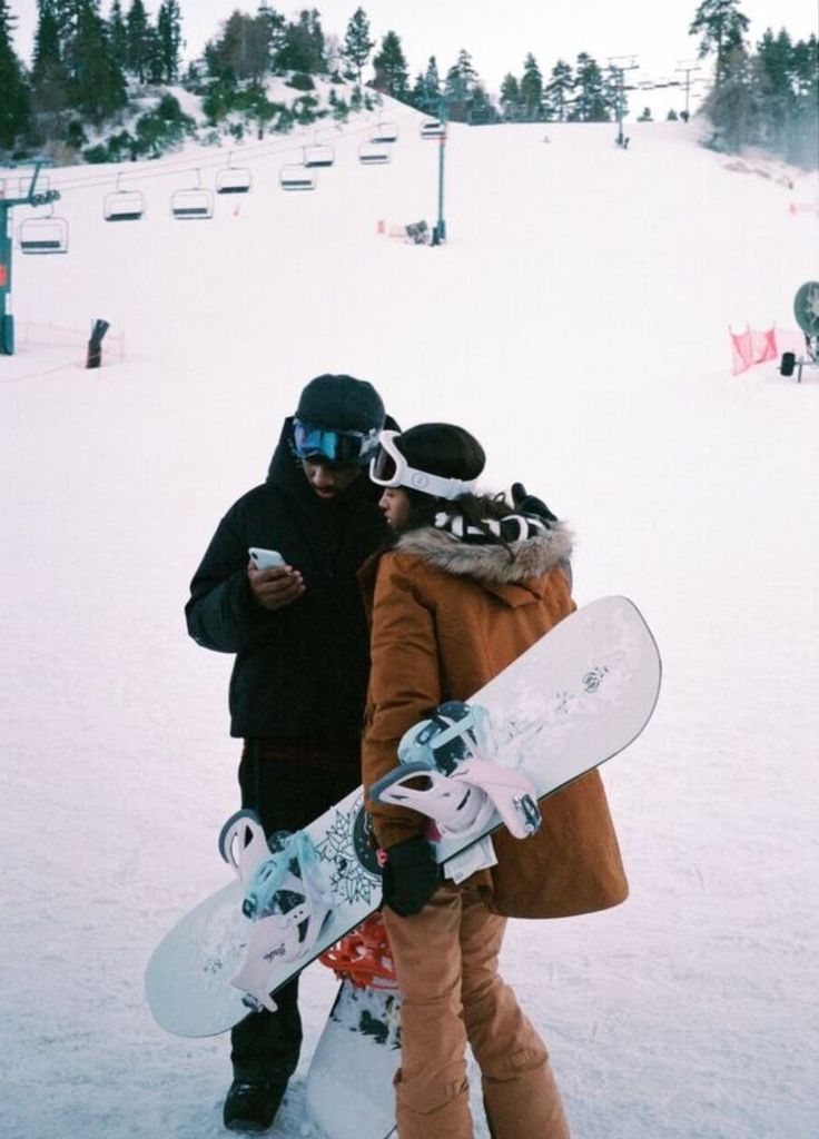 two snowboarders standing in the snow with their boards attached to their shoulders and looking at something