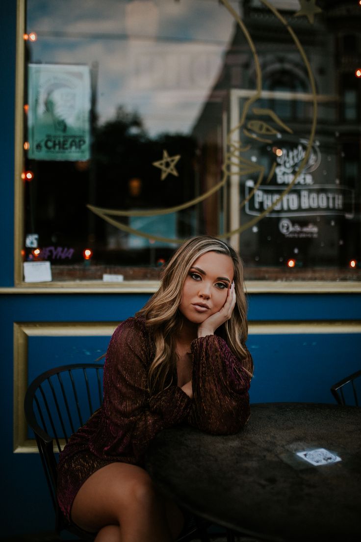 a beautiful woman sitting at a table in front of a store window with her hand on her chin