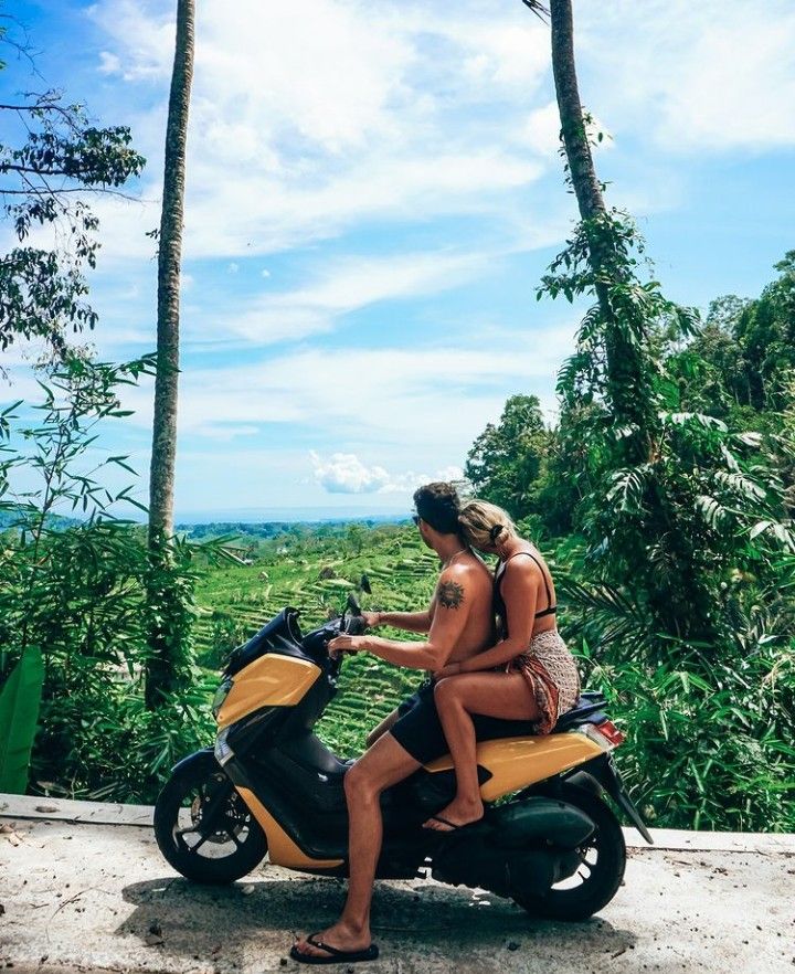 two people sitting on a scooter in front of some palm trees and the ocean