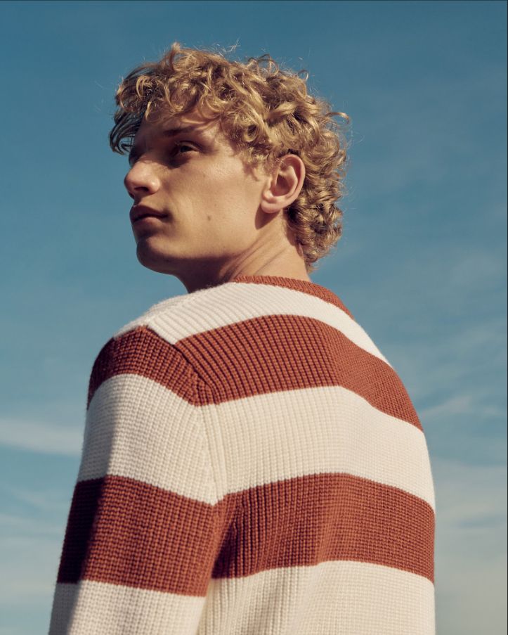 a young man with curly hair wearing a striped sweater looking off to the side against a blue sky