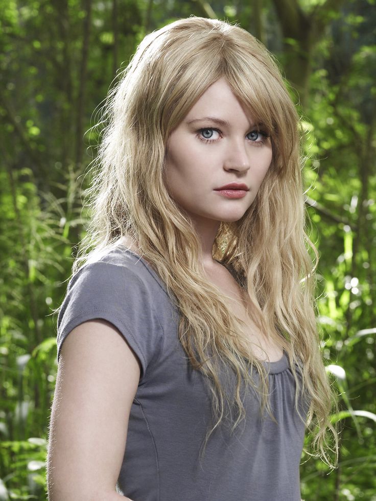 a woman with long blonde hair standing in front of some trees and bushes, looking at the camera