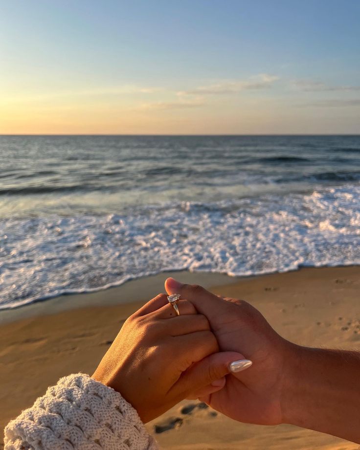 two people holding hands on the beach with waves coming in to shore and one person wearing a ring